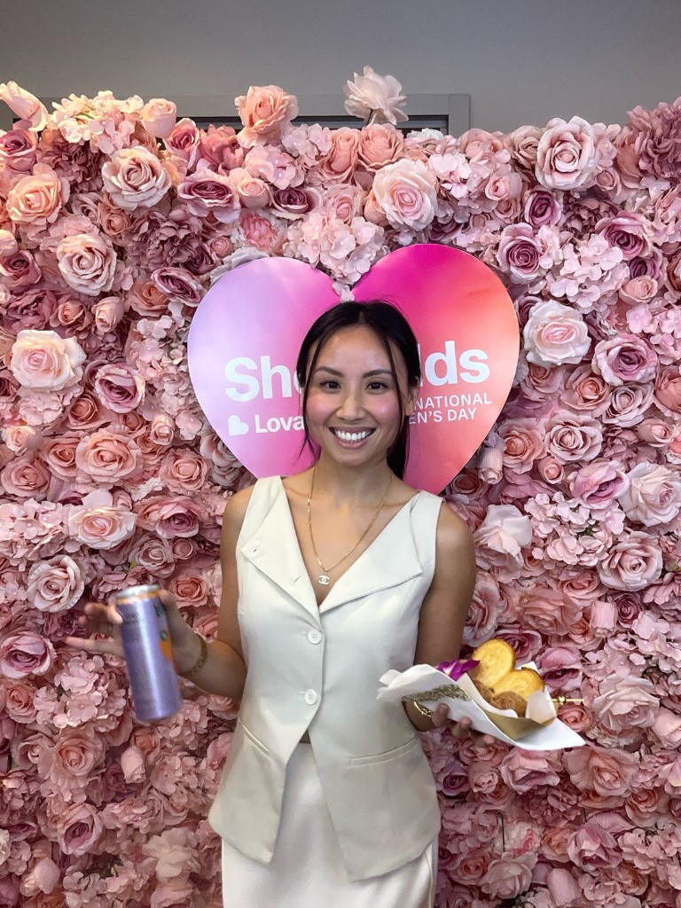Woman smiling in front of pink floral wall with She Builds and International Women's Day signs at Plug and Play event